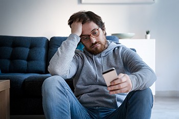 How to Watch For Signs of Relapse During the Coronavirus Isolation, man wearing glasses in a hoodie looking at his phone on the floor.