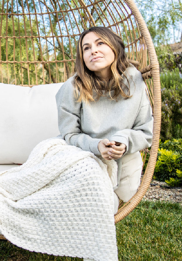 Woman with brown hair sitting outside in a chair with a knit blanket on.