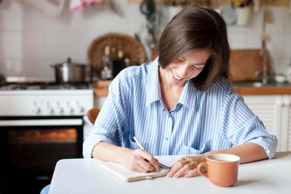 Beating addiction in the new year, woman in a blue shirt smiling while she journals in the kitchen.