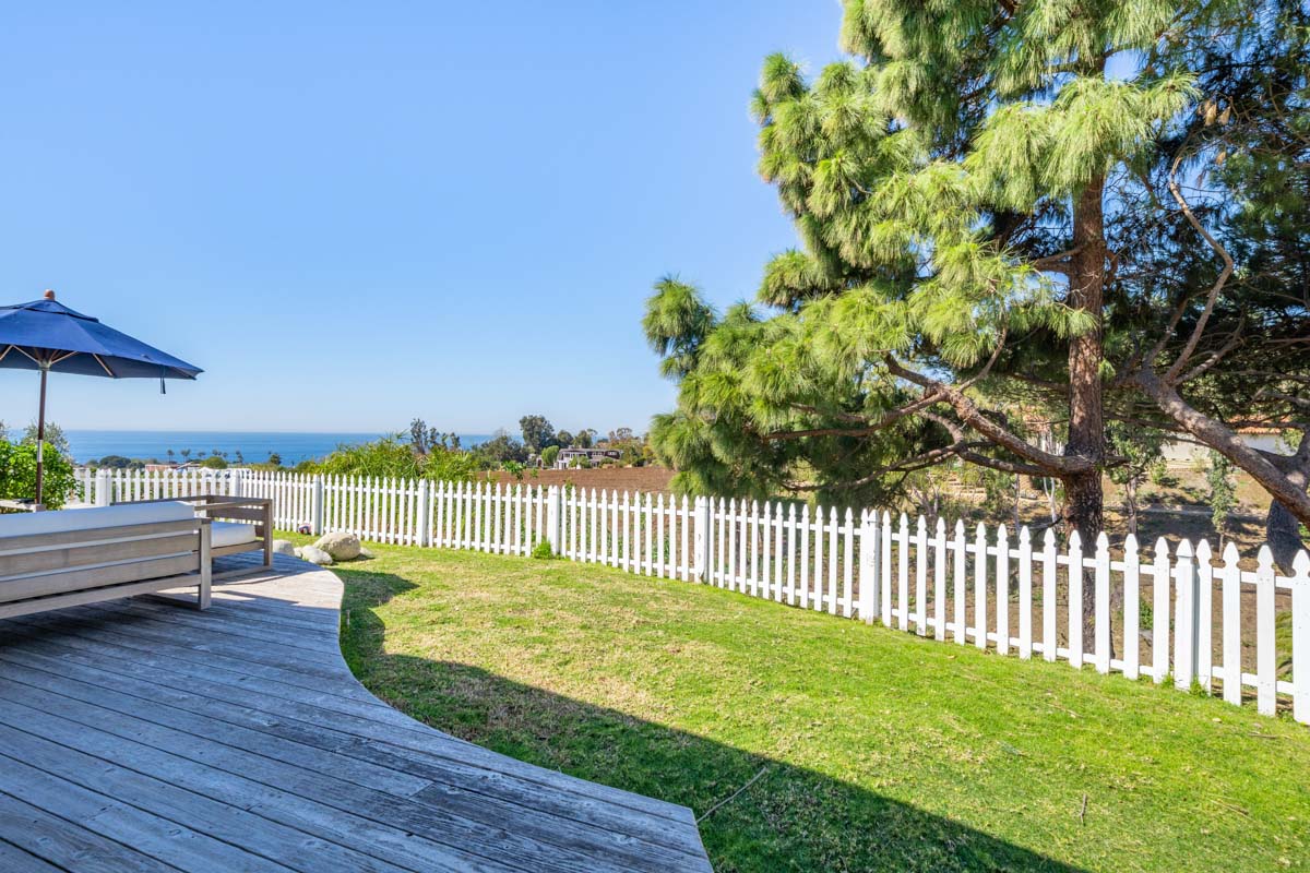 Backyard view at Cliffside Malibu, a residential treatment center in Malibu, California. Wooden deck by the large grass yard surrounded by a white picket fence. Blue umbrella and view of the ocean in the back.