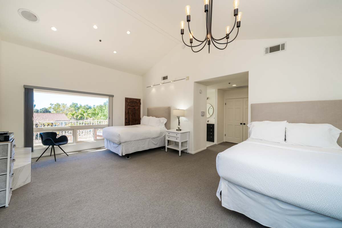 Bedroom with two beds at Cliffside Malibu, a residential treatment center in Malibu California. Two beds in a carpeted room, white comforters over the beds and a large open window in the left corner.