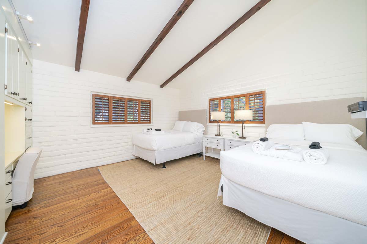 Two beds in a bedroom over wood floors at Cliffside Malibu, a residential treatment center in Malibu, California. Exposed wooden beams on ceiling and white brick walls.