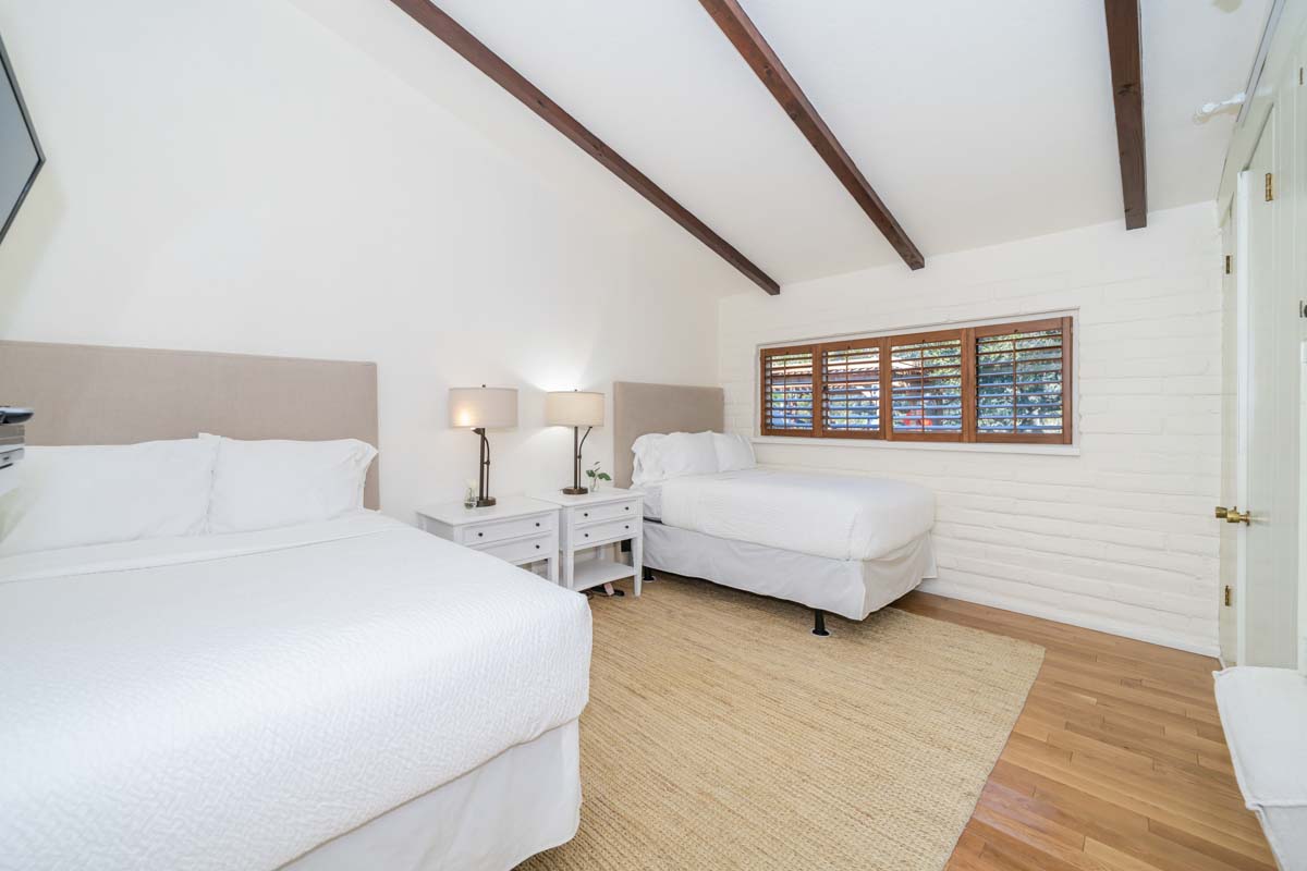 Two bed, bedroom at Cliffside Malibu, a residential treatment center in Malibu, California. Beds have white comforters on them, are atop a brown textured rug and have two white bedside tables with lamps on top of them.