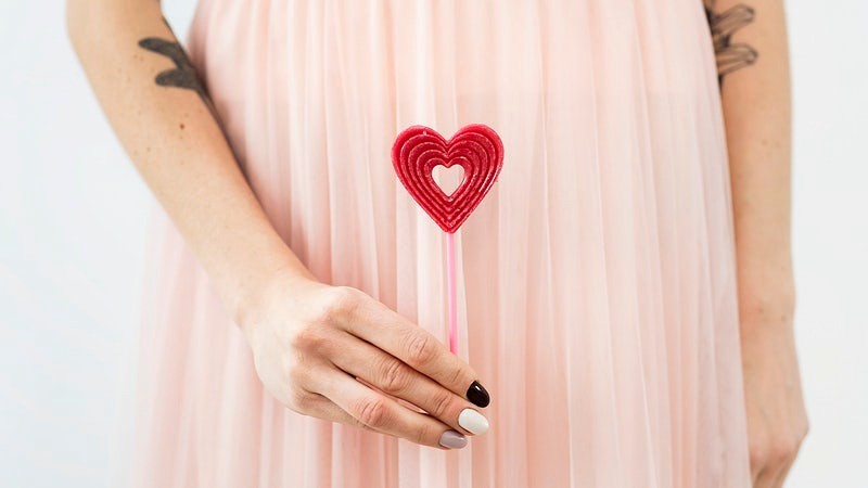 Woman in a pink dress with tattoo's holding a red heart lollipop, surviving Valentine’s Day in recovery.