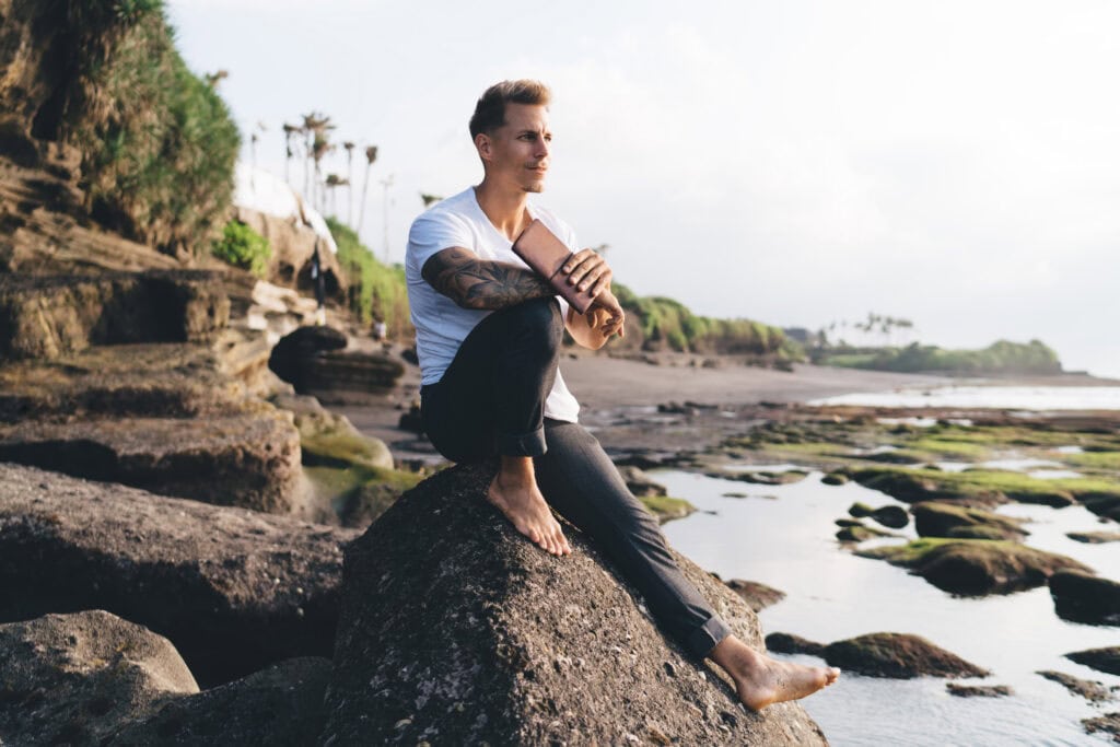 Full body side view of bearded male in casual clothes holding book while sitting on rocky coast of river and looking at camera on sunny day